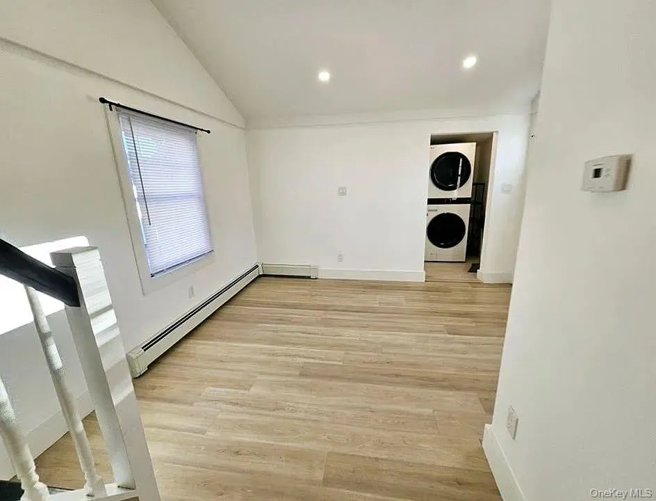 Washroom featuring baseboard heating, light wood-type flooring, stacked washer / drying machine, recessed lighting, and vaulted ceiling Washroom featuring baseboard heating, light wood-type flooring, stacked washer / drying machine, recessed lighting, and vaulted ceiling