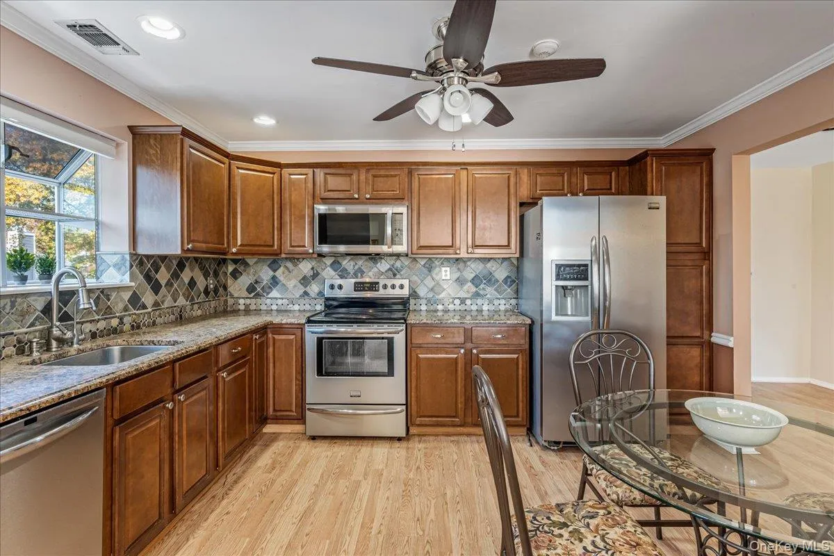 Kitchen featuring stainless steel appliances, light stone countertops, crown molding, decorative backsplash, and brown cabinetry Kitchen featuring stainless steel appliances, light stone countertops, crown molding, decorative backsplash, and brown cabinetry