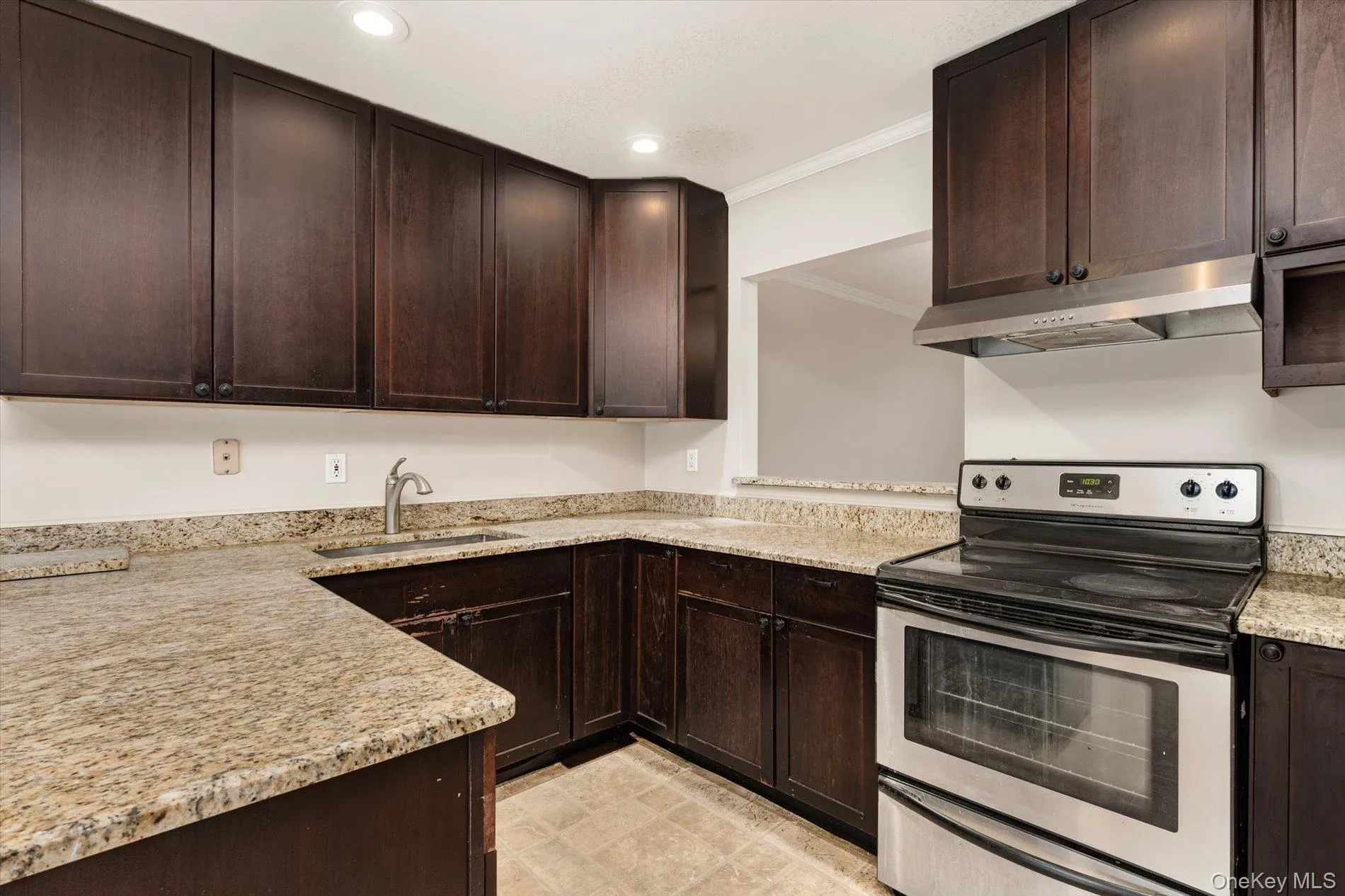Kitchen with dark brown cabinets, stainless steel electric range oven, ornamental molding, under cabinet range hood, and light stone counters Kitchen with dark brown cabinets, stainless steel electric range oven, ornamental molding, under cabinet range hood, and light stone counters