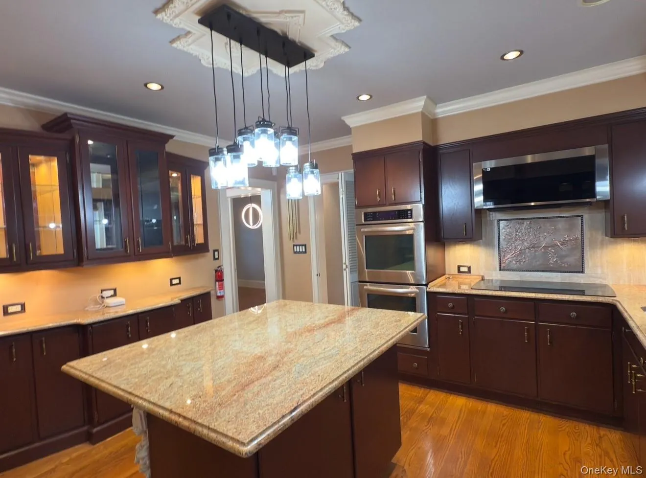 Kitchen featuring ornamental molding, a kitchen island, light wood-type flooring, stainless steel appliances, and decorative light fixtures Kitchen featuring ornamental molding, a kitchen island, light wood-type flooring, stainless steel appliances, and decorative light fixtures