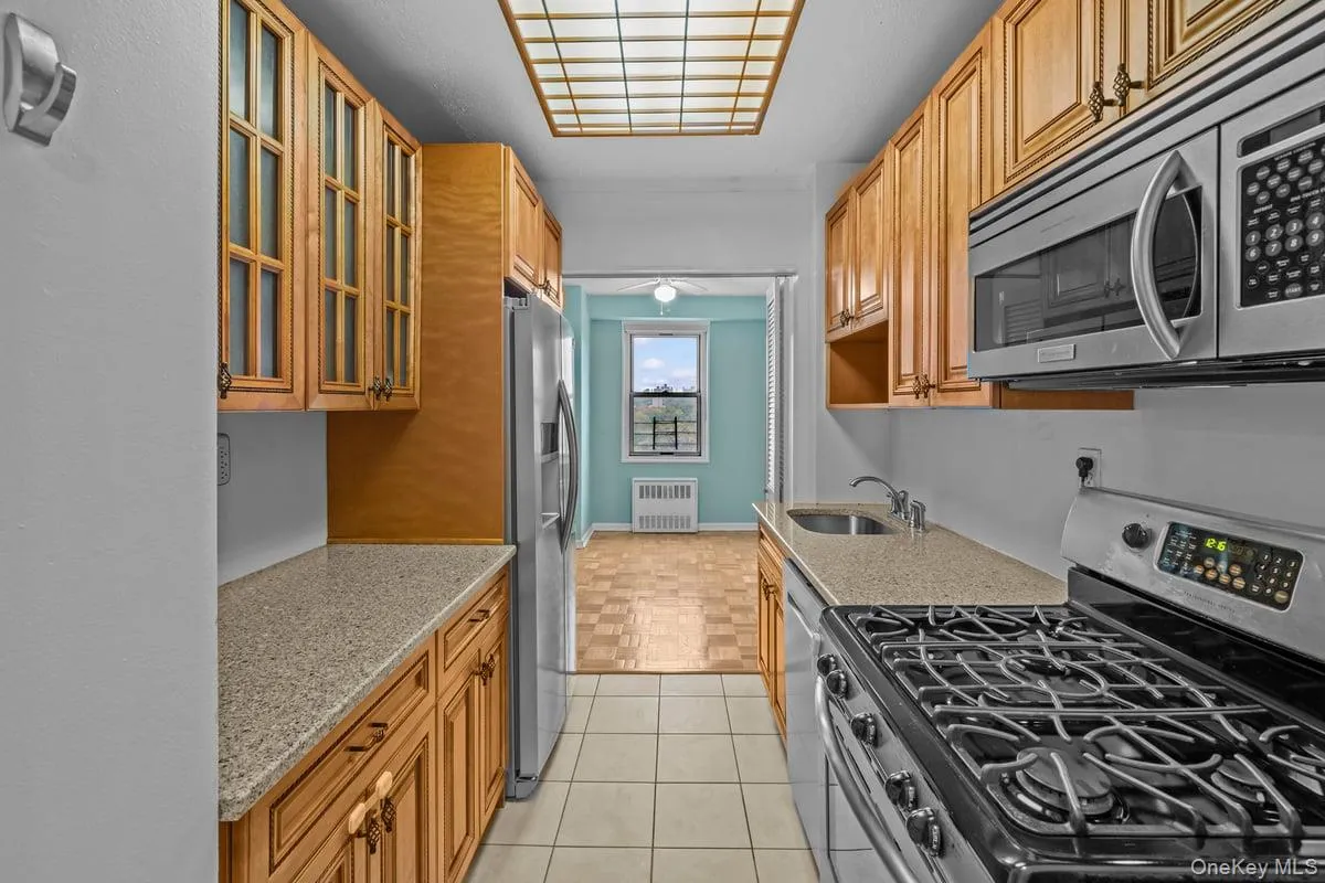 Kitchen featuring appliances with stainless steel finishes, glass insert cabinets, light stone counters, radiator, and brown cabinets Kitchen featuring appliances with stainless steel finishes, glass insert cabinets, light stone counters, radiator, and brown cabinets