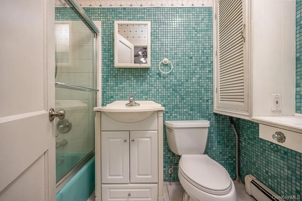 Full bathroom with vanity, bath / shower combo with glass door, a baseboard heating unit, tile walls, and tile patterned floors Full bathroom with vanity, bath / shower combo with glass door, a baseboard heating unit, tile walls, and tile patterned floors