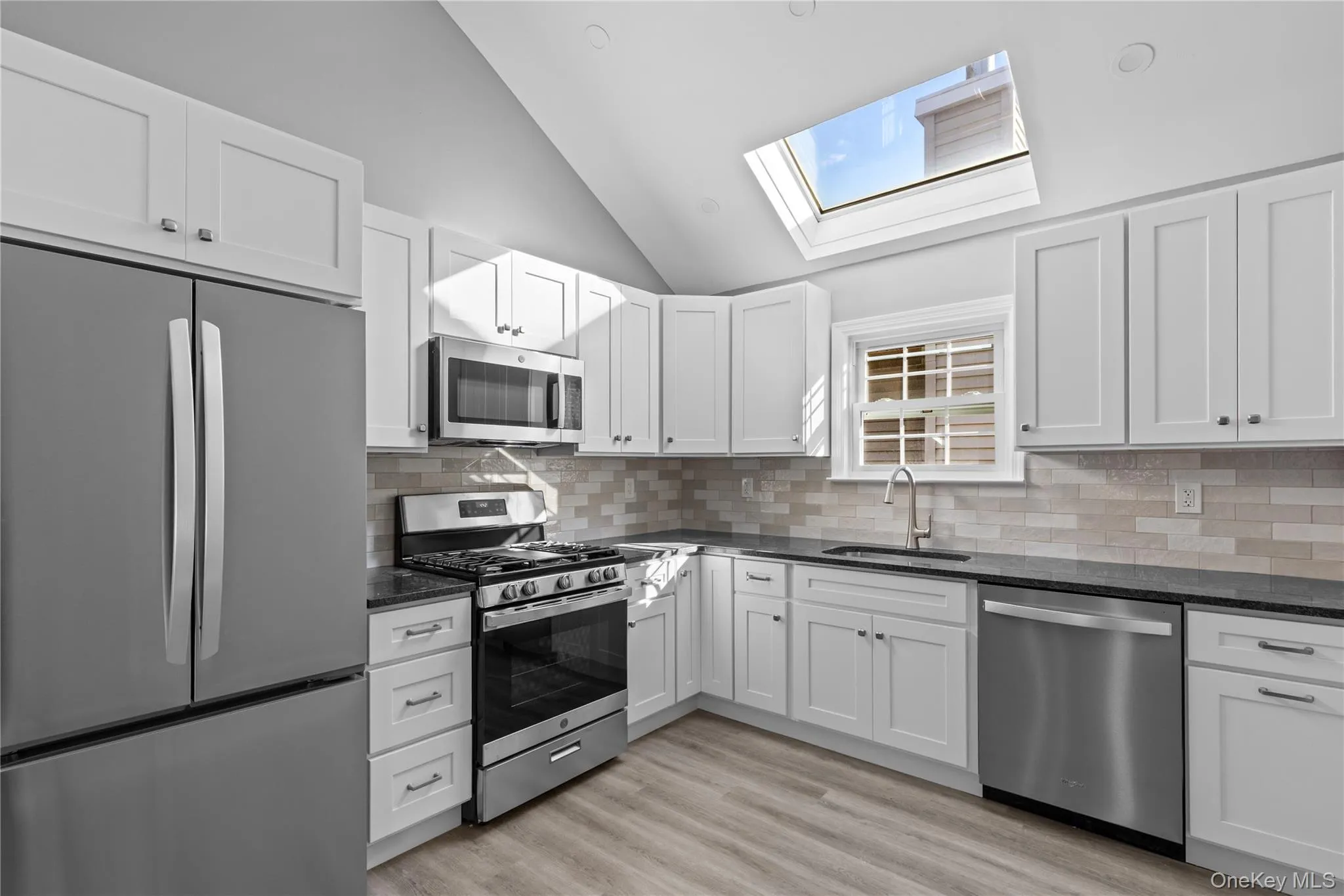 Kitchen with stainless steel appliances, white cabinets, dark stone counters, backsplash, and high vaulted ceiling Kitchen with stainless steel appliances, white cabinets, dark stone counters, backsplash, and high vaulted ceiling