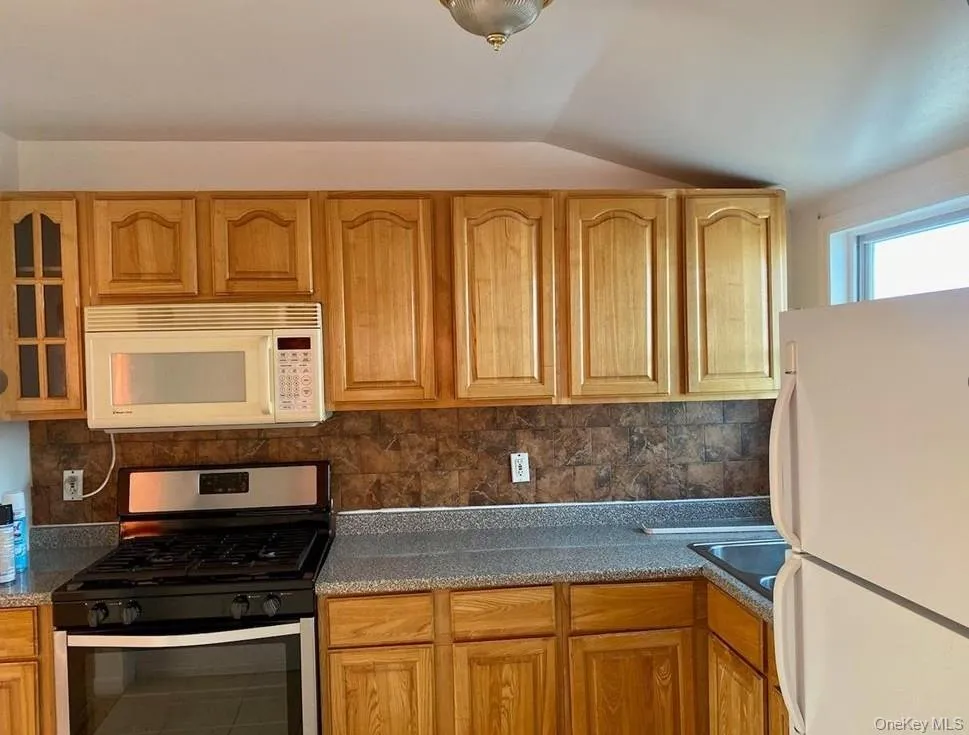 Kitchen featuring white appliances, decorative backsplash, vaulted ceiling, and brown cabinetry Kitchen featuring white appliances, decorative backsplash, vaulted ceiling, and brown cabinetry