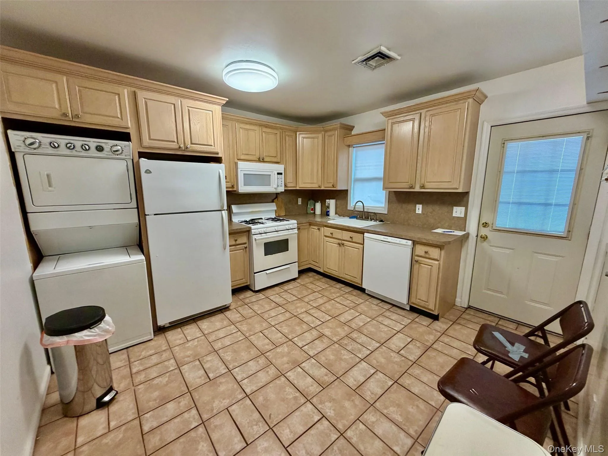 Kitchen with light brown cabinets, white appliances, stacked washer / dryer, light countertops, and light tile patterned flooring Kitchen with light brown cabinets, white appliances, stacked washer / dryer, light countertops, and light tile patterned flooring