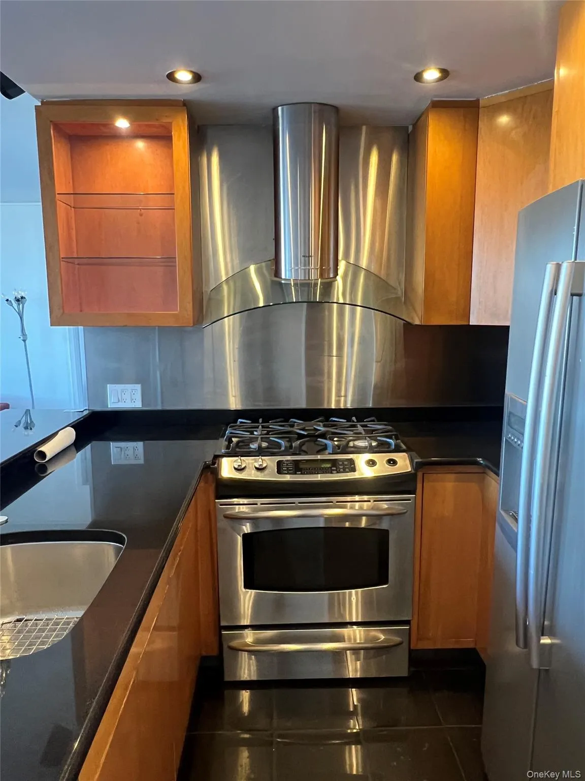 Kitchen featuring appliances with stainless steel finishes, wall chimney range hood, brown cabinets, dark tile patterned flooring, and recessed lighting Kitchen featuring appliances with stainless steel finishes, wall chimney range hood, brown cabinets, dark tile patterned flooring, and recessed lighting