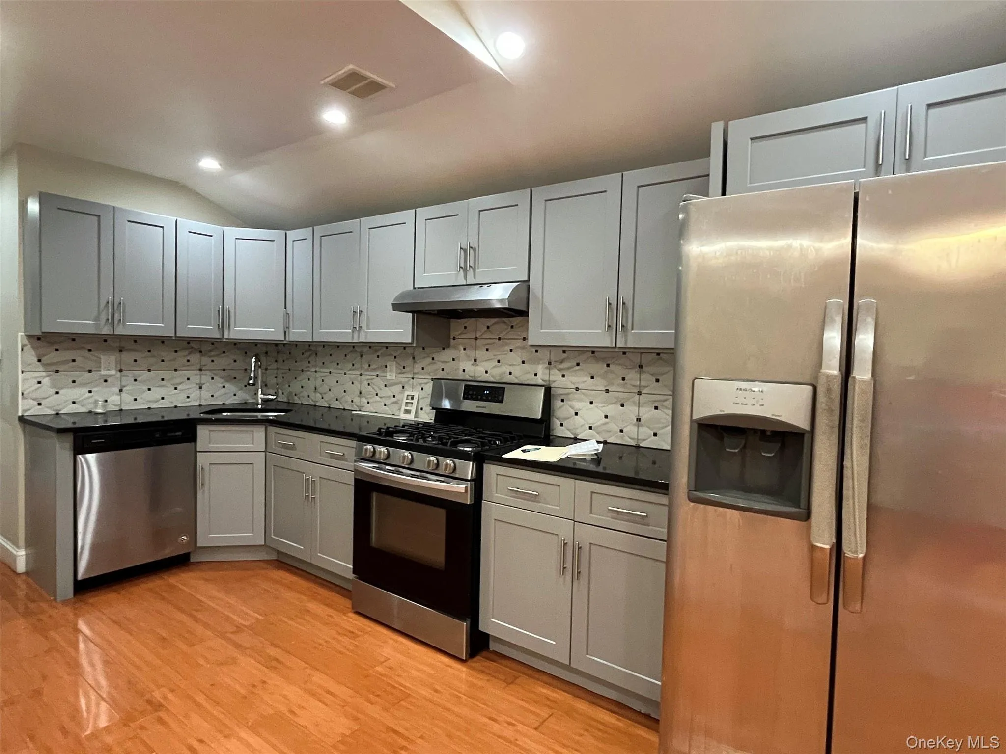 Kitchen with appliances with stainless steel finishes, gray cabinets, decorative backsplash, light wood-style flooring, and lofted ceiling Kitchen with appliances with stainless steel finishes, gray cabinets, decorative backsplash, light wood-style flooring, and lofted ceiling