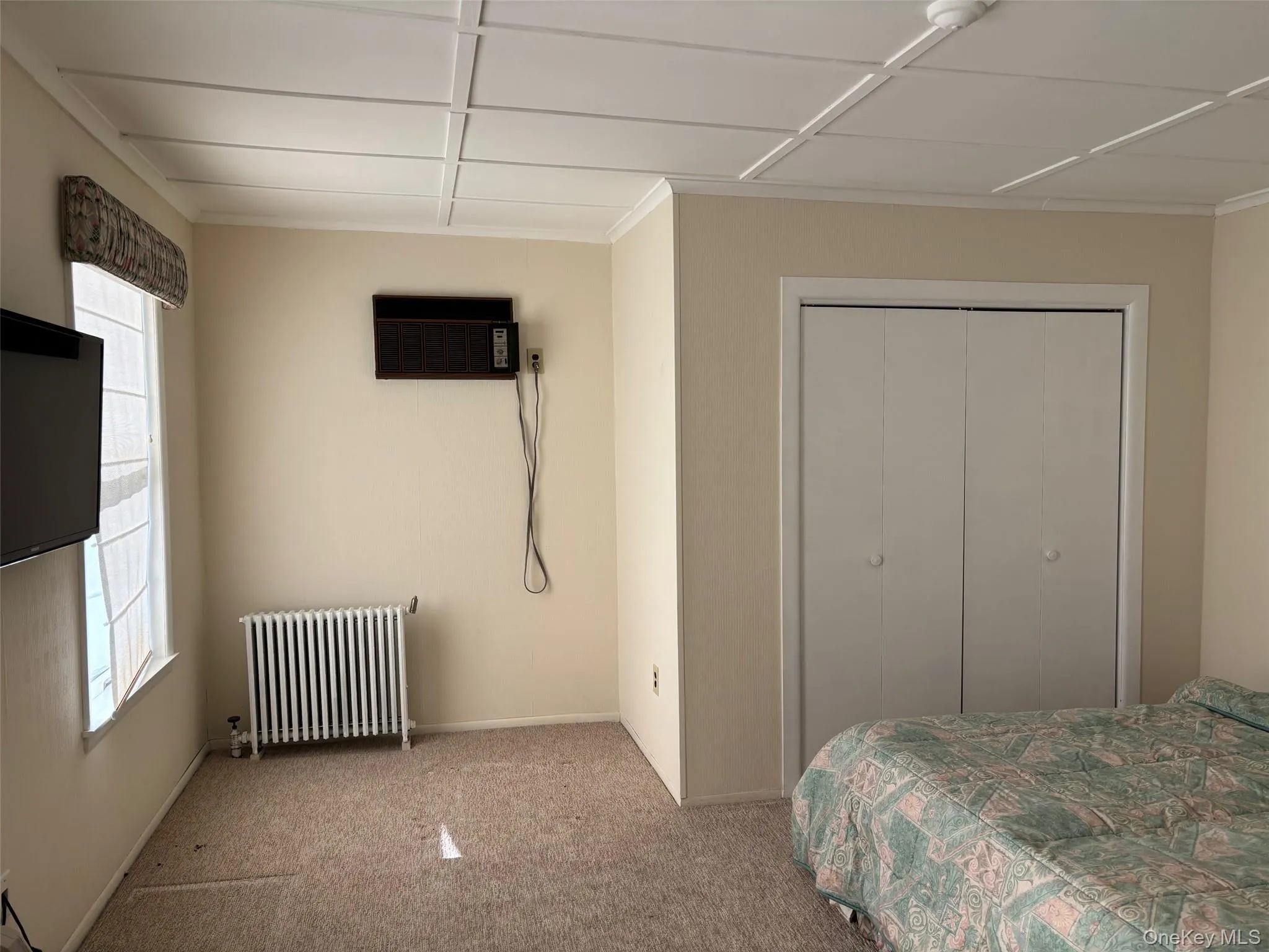 Carpeted bedroom featuring radiator heating unit, a closet, and a wall mounted air conditioner Carpeted bedroom featuring radiator heating unit, a closet, and a wall mounted air conditioner