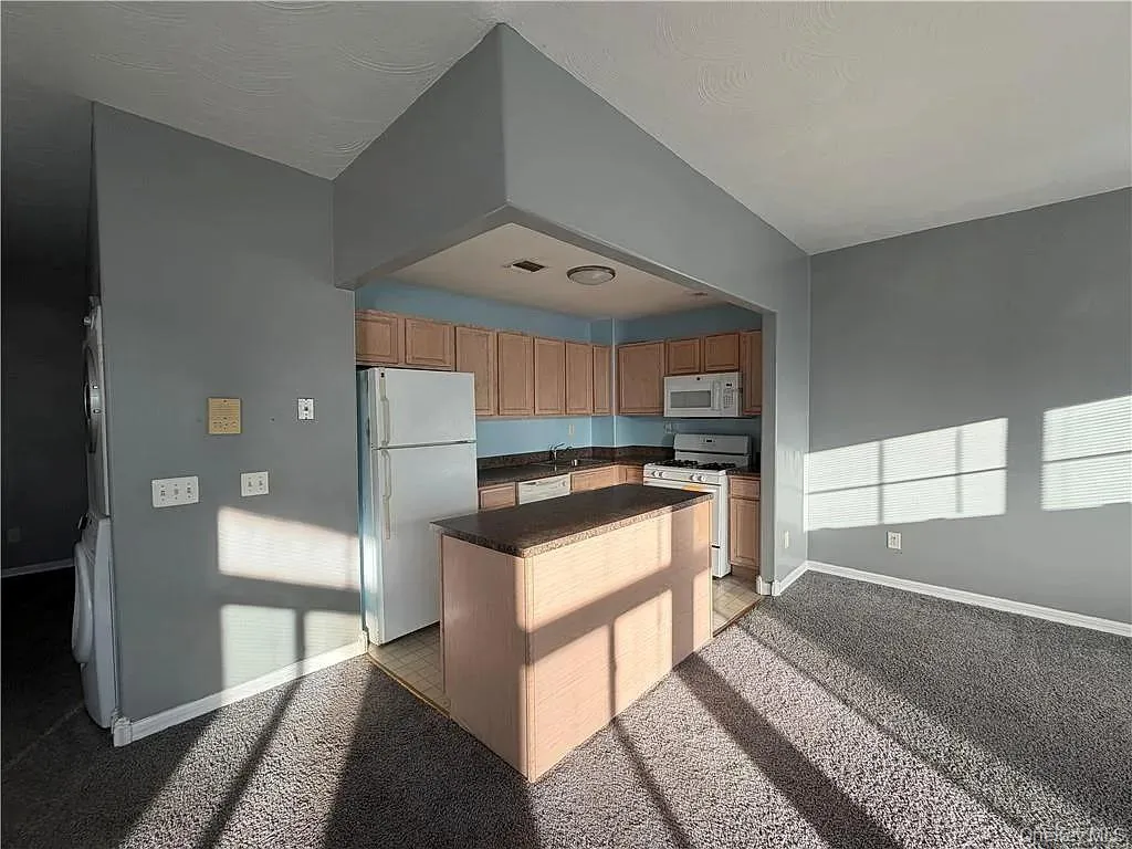 Kitchen featuring white appliances, a center island, dark countertops, and light colored carpet Kitchen featuring white appliances, a center island, dark countertops, and light colored carpet
