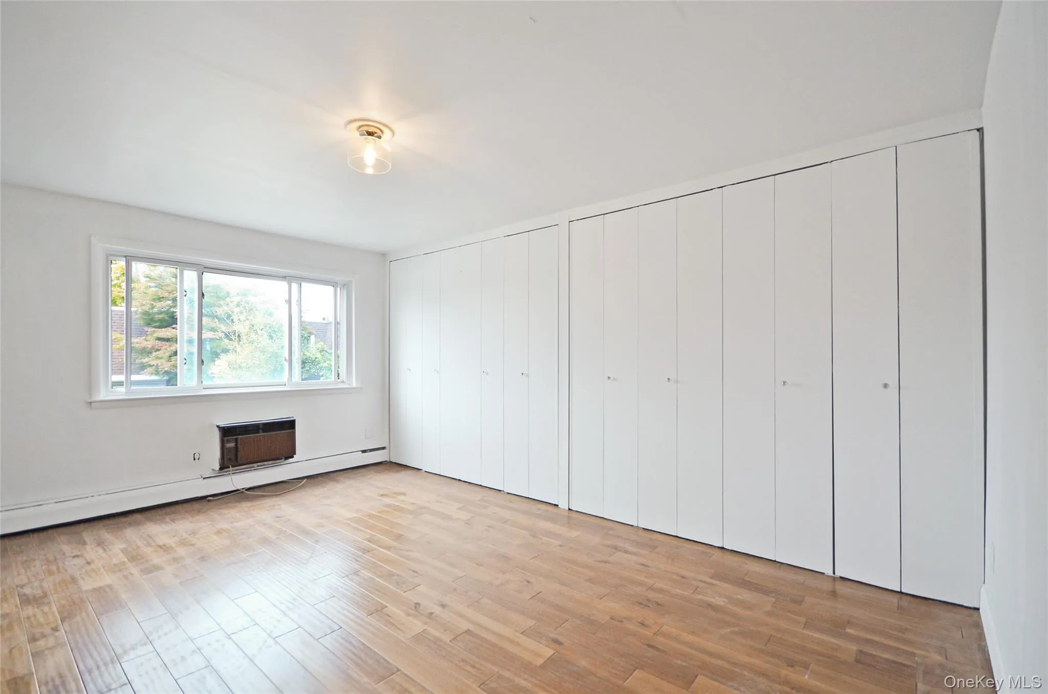 Unfurnished bedroom featuring two closets, wood finished floors, baseboard heating, and a wall unit AC Unfurnished bedroom featuring two closets, wood finished floors, baseboard heating, and a wall unit AC