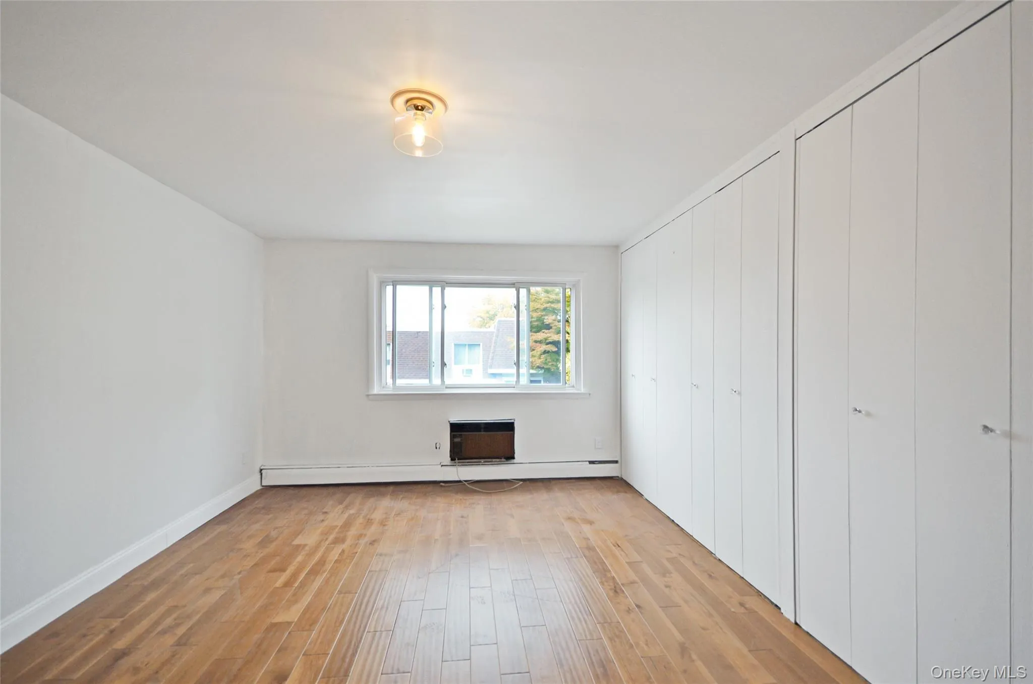Unfurnished bedroom featuring light wood-style floors, multiple closets, a baseboard heating unit, and an AC wall unit Unfurnished bedroom featuring light wood-style floors, multiple closets, a baseboard heating unit, and an AC wall unit
