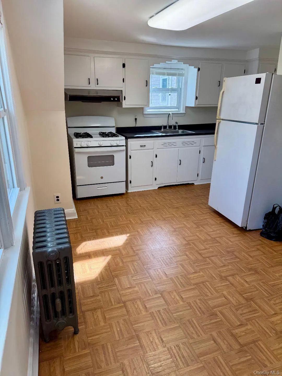 Kitchen with white appliances, dark countertops, white cabinetry, under cabinet range hood, and radiator Kitchen with white appliances, dark countertops, white cabinetry, under cabinet range hood, and radiator