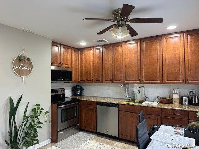 Kitchen with appliances with stainless steel finishes, decorative backsplash, light stone countertops, ceiling fan, and brown cabinetry Kitchen with appliances with stainless steel finishes, decorative backsplash, light stone countertops, ceiling fan, and brown cabinetry