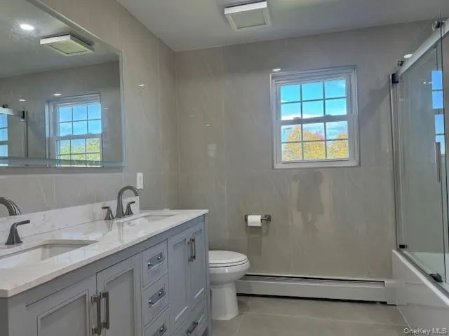 Full bath featuring double vanity, combined bath / shower with glass door, light tile patterned floors, a baseboard radiator, and tile walls Full bath featuring double vanity, combined bath / shower with glass door, light tile patterned floors, a baseboard radiator, and tile walls