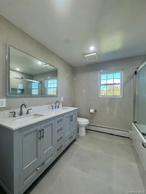 Full bath with double vanity, a baseboard radiator, shower / bath combination with glass door, tile walls, and light tile patterned floors Full bath with double vanity, a baseboard radiator, shower / bath combination with glass door, tile walls, and light tile patterned floors