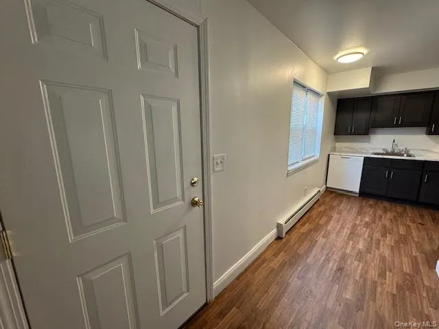 Kitchen with light countertops, a baseboard heating unit, dark wood-style flooring, white dishwasher, and dark cabinetry Kitchen with light countertops, a baseboard heating unit, dark wood-style flooring, white dishwasher, and dark cabinetry