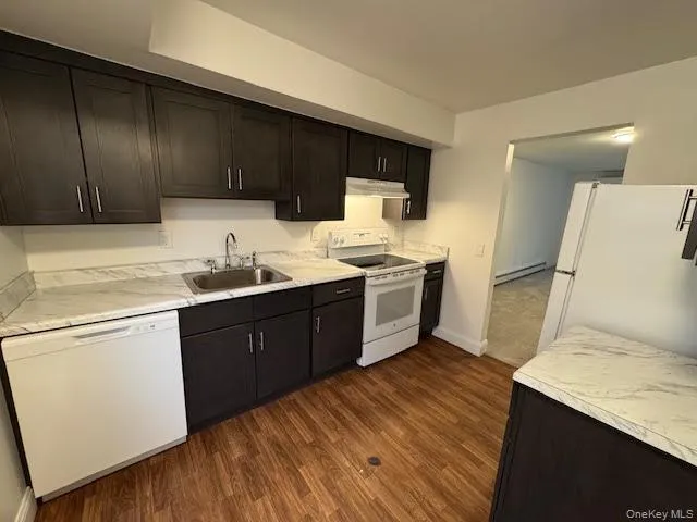Kitchen with white appliances, light countertops, dark wood-style flooring, under cabinet range hood, and baseboard heating Kitchen with white appliances, light countertops, dark wood-style flooring, under cabinet range hood, and baseboard heating