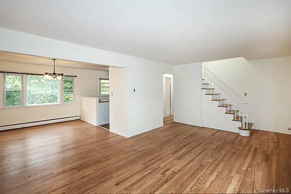 Unfurnished living room with a chandelier, light wood finished floors, stairs, and a baseboard heating unit Unfurnished living room with a chandelier, light wood finished floors, stairs, and a baseboard heating unit