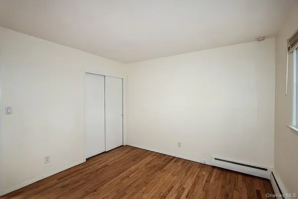 Unfurnished bedroom with dark wood finished floors, a baseboard radiator, a closet, and multiple windows Unfurnished bedroom with dark wood finished floors, a baseboard radiator, a closet, and multiple windows