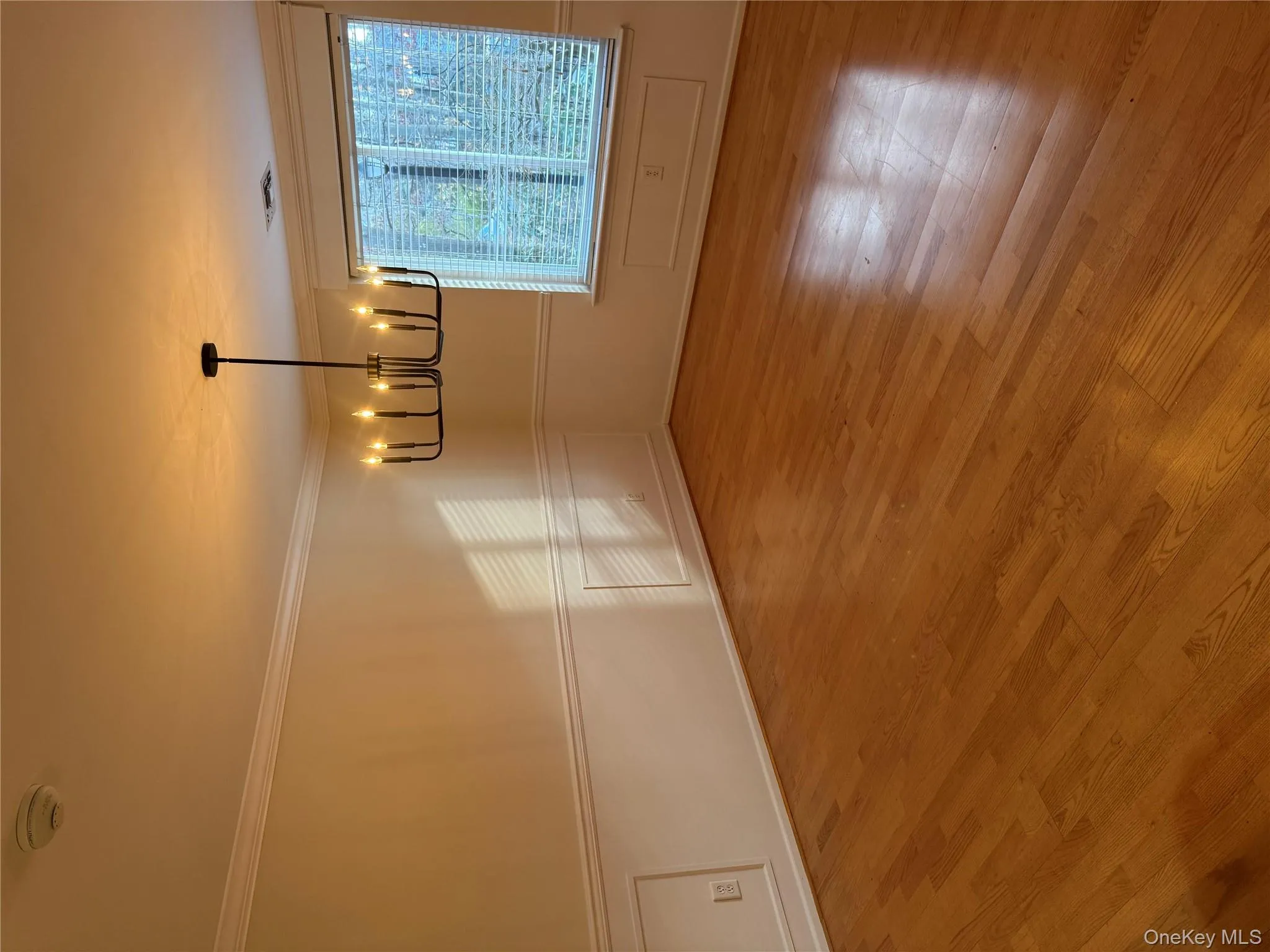 Empty room with light wood-type flooring, crown molding, a wainscoted wall, a chandelier, and a smoke detector Empty room with light wood-type flooring, crown molding, a wainscoted wall, a chandelier, and a smoke detector