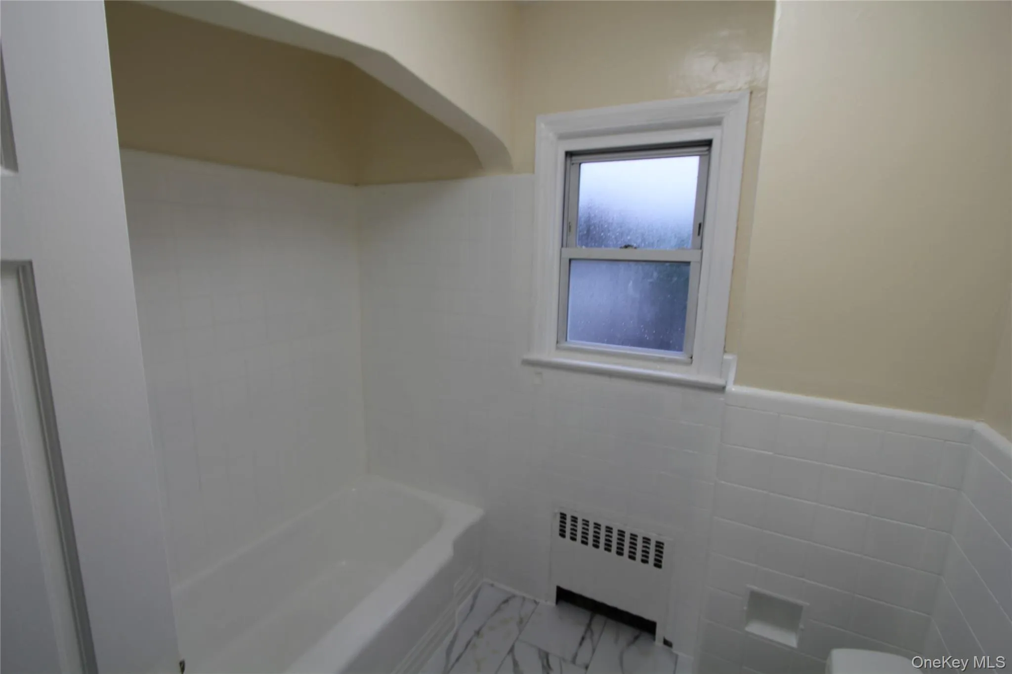 Full bathroom with tile walls, radiator heating unit, wainscoting, light marble finish flooring, and washtub / shower combination Full bathroom with tile walls, radiator heating unit, wainscoting, light marble finish flooring, and washtub / shower combination