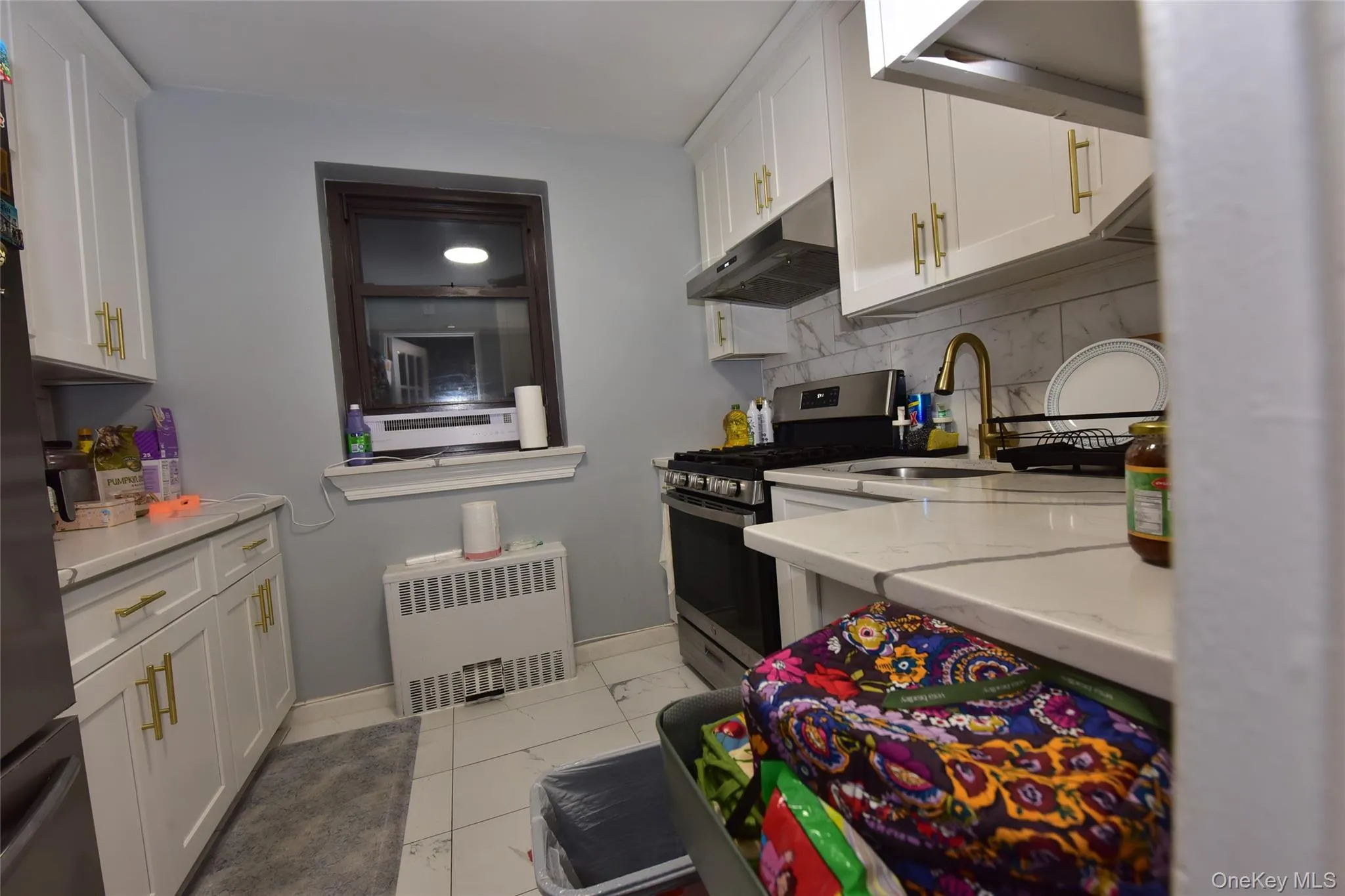 Kitchen featuring white cabinetry, stainless steel appliances, under cabinet range hood, radiator heating unit, and light stone countertops Kitchen featuring white cabinetry, stainless steel appliances, under cabinet range hood, radiator heating unit, and light stone countertops