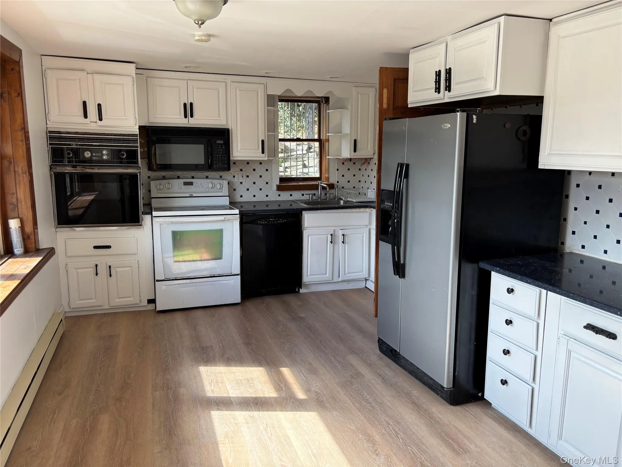 Kitchen with decorative backsplash, white cabinets, black appliances, a baseboard radiator, and light wood-type flooring Kitchen with decorative backsplash, white cabinets, black appliances, a baseboard radiator, and light wood-type flooring