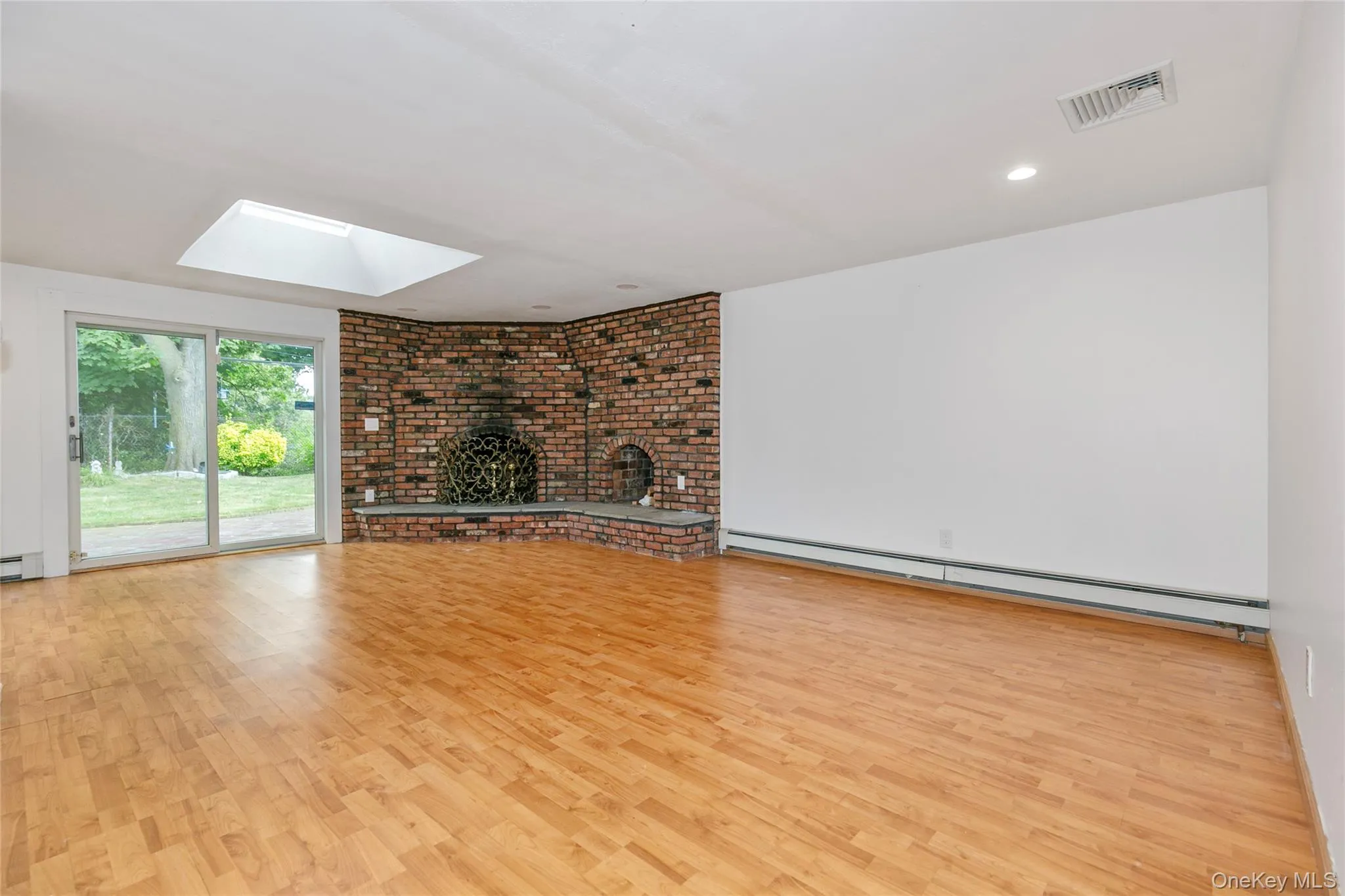 Unfurnished living room featuring a baseboard heating unit, light wood-style flooring, a skylight, a fireplace, and brick wall Unfurnished living room featuring a baseboard heating unit, light wood-style flooring, a skylight, a fireplace, and brick wall