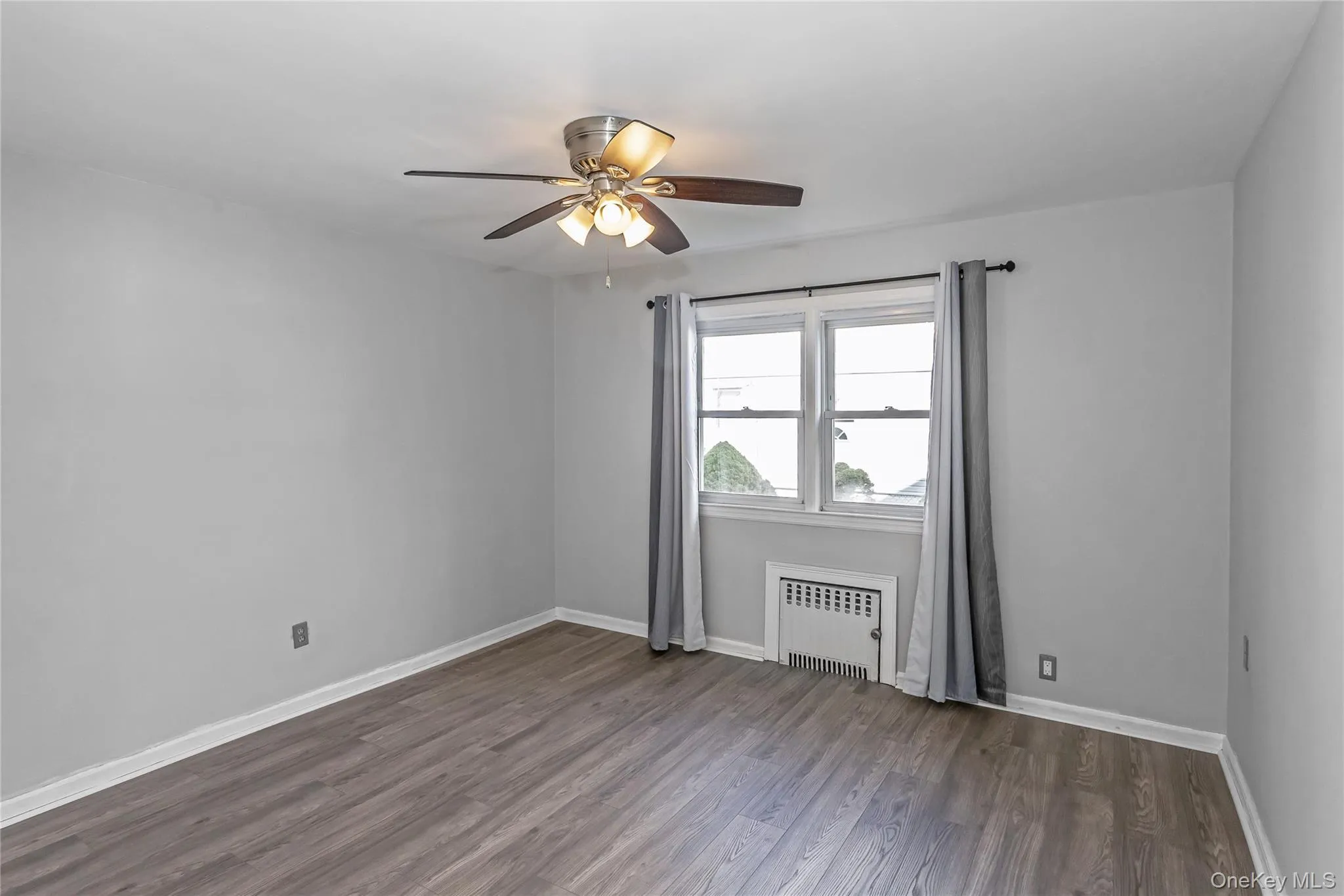 Empty room with dark wood-type flooring, baseboards, ceiling fan, and radiator heating unit Empty room with dark wood-type flooring, baseboards, ceiling fan, and radiator heating unit