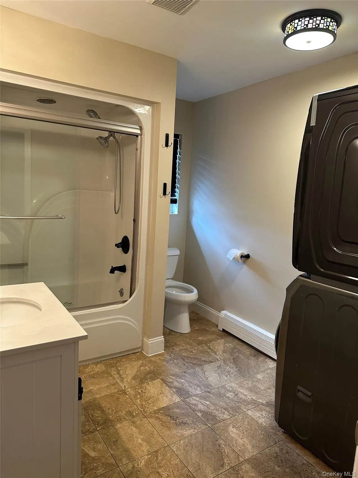 Bathroom with shower / bath combination with glass door, baseboard heating, vanity, light stone finish flooring, and stacked washer / dryer Bathroom with shower / bath combination with glass door, baseboard heating, vanity, light stone finish flooring, and stacked washer / dryer