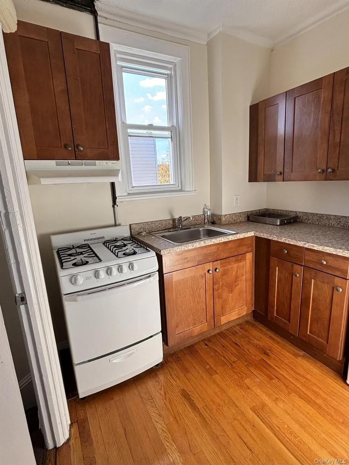 Kitchen featuring white range with gas stovetop, light wood finished floors, crown molding, under cabinet range hood, and brown cabinetry Kitchen featuring white range with gas stovetop, light wood finished floors, crown molding, under cabinet range hood, and brown cabinetry