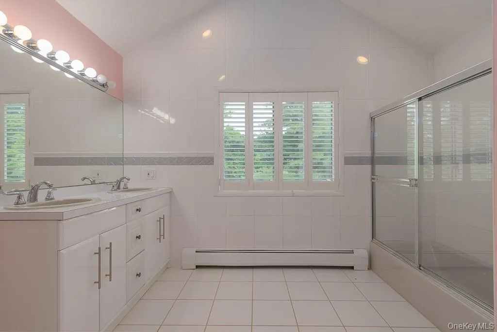 Full bath with tile walls, light tile patterned floors, double vanity, baseboard heating, and enclosed tub / shower combo Full bath with tile walls, light tile patterned floors, double vanity, baseboard heating, and enclosed tub / shower combo