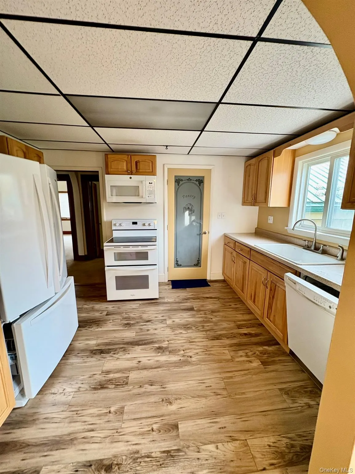 Kitchen with white appliances, light countertops, light wood finished floors, and a paneled ceiling Kitchen with white appliances, light countertops, light wood finished floors, and a paneled ceiling