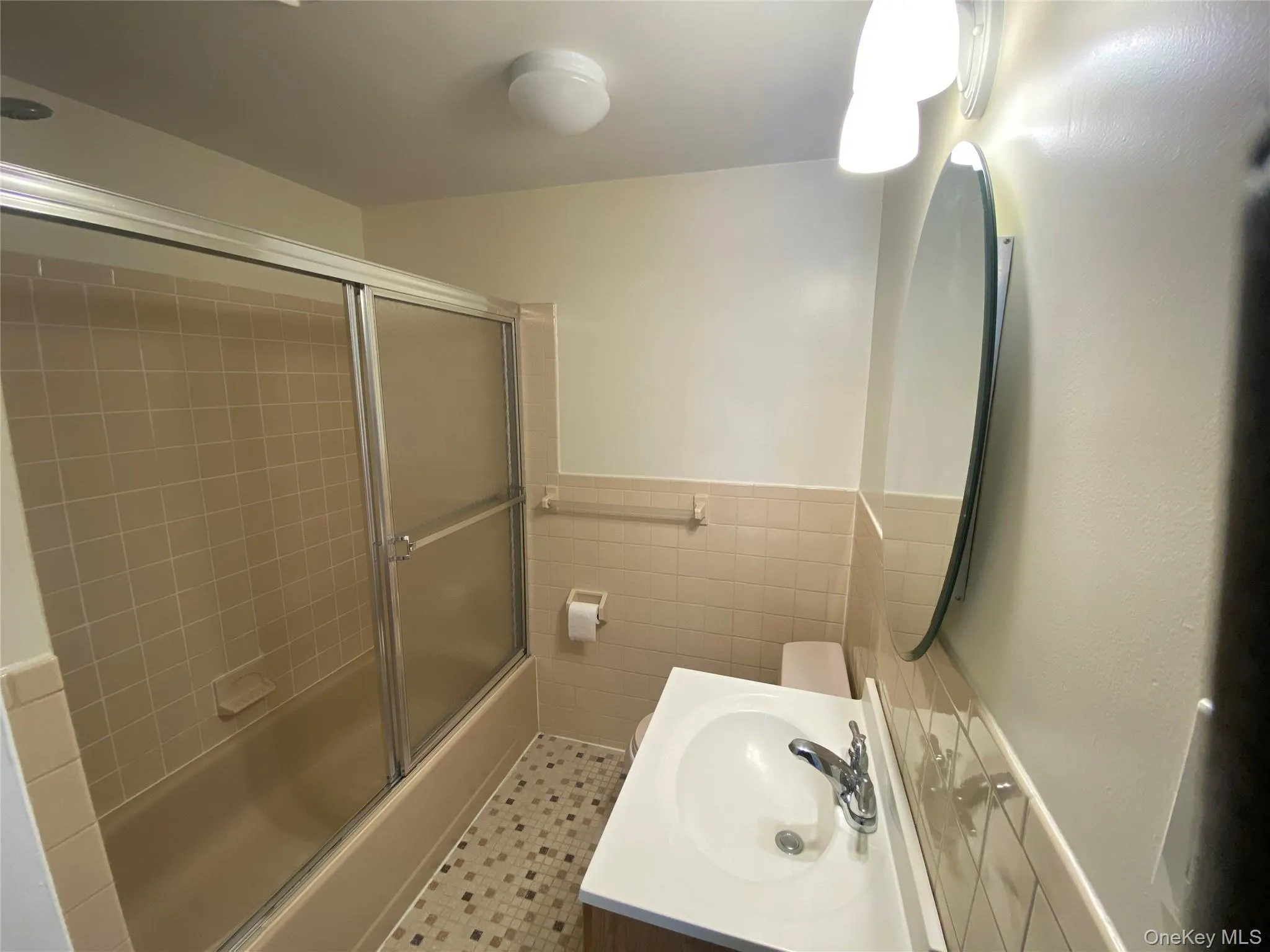 Full bathroom with tile walls, vanity, bath / shower combo with glass door, wainscoting, and light tile patterned flooring Full bathroom with tile walls, vanity, bath / shower combo with glass door, wainscoting, and light tile patterned flooring