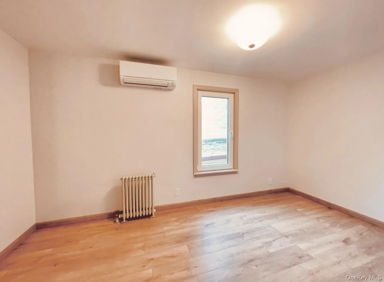Empty room with radiator heating unit, light wood-style flooring, and a wall mounted AC Empty room with radiator heating unit, light wood-style flooring, and a wall mounted AC