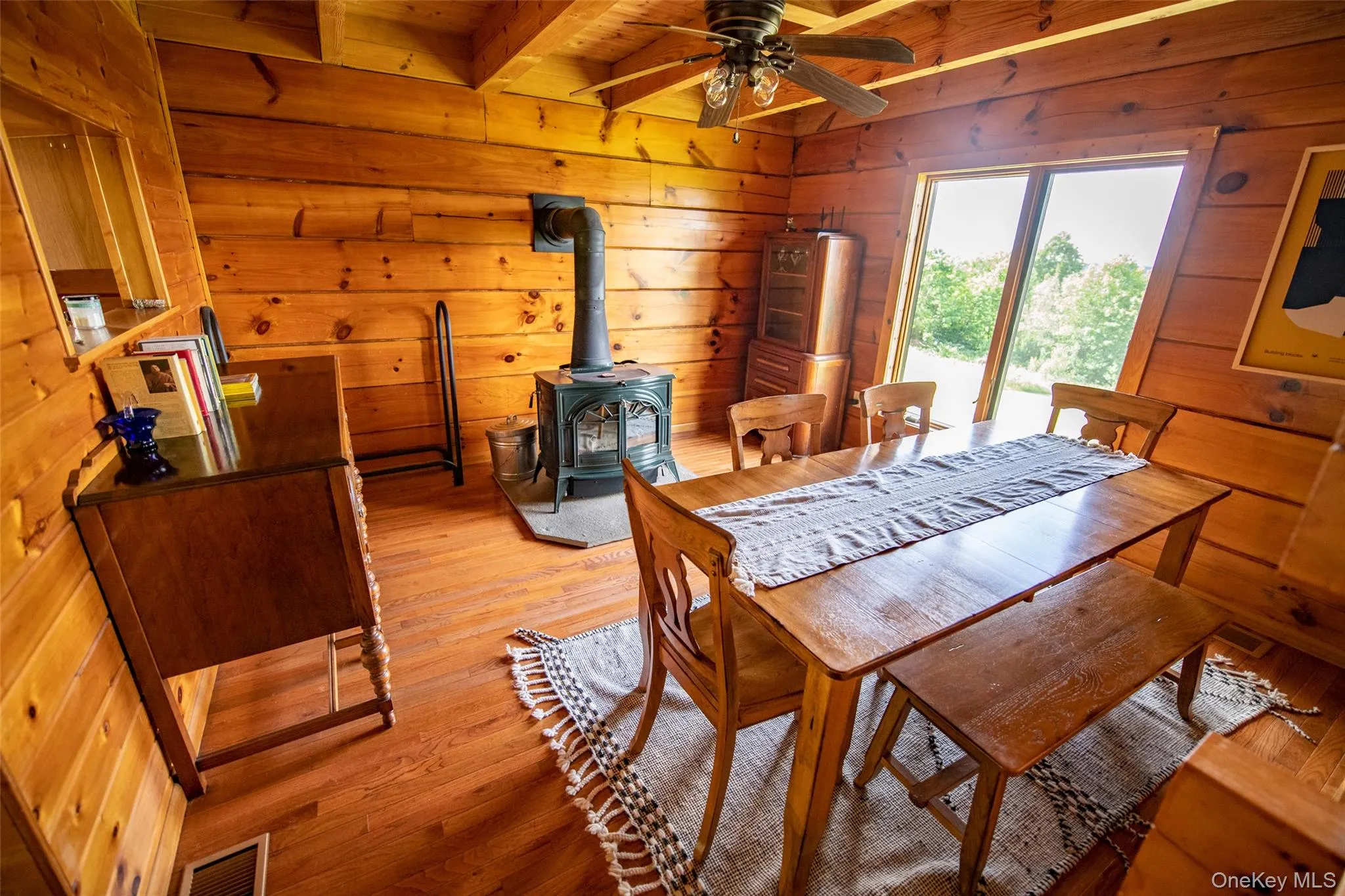 Dining room with a wood stove, wood-type flooring, wooden walls, ceiling fan, and a wood ceiling with exposed beams Dining room with a wood stove, wood-type flooring, wooden walls, ceiling fan, and a wood ceiling with exposed beams