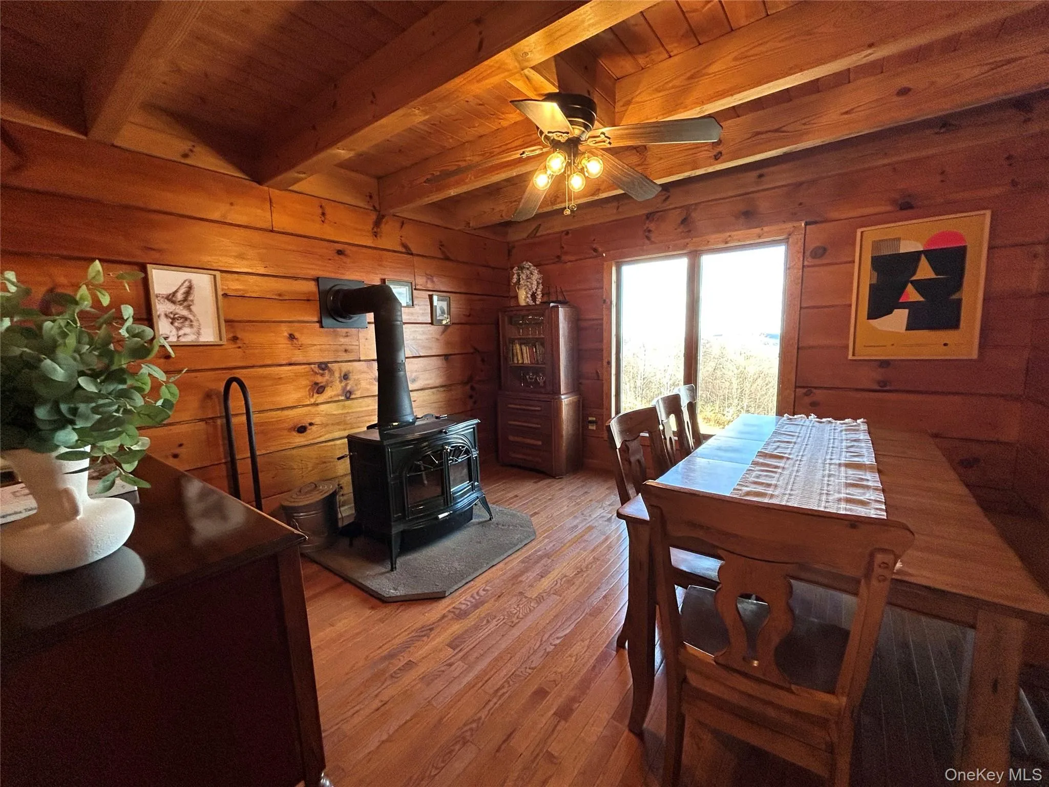 Dining room featuring a wood stove, wood finished floors, a wooden ceiling with exposed beams, wood walls, and a ceiling fan Dining room featuring a wood stove, wood finished floors, a wooden ceiling with exposed beams, wood walls, and a ceiling fan