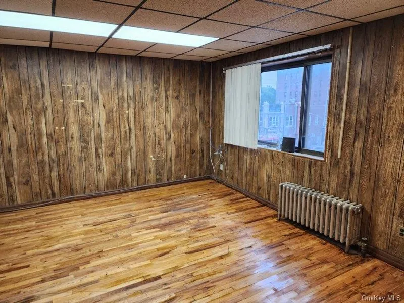 Spare room with wood walls, radiator, light wood-style flooring, and a paneled ceiling Spare room with wood walls, radiator, light wood-style flooring, and a paneled ceiling