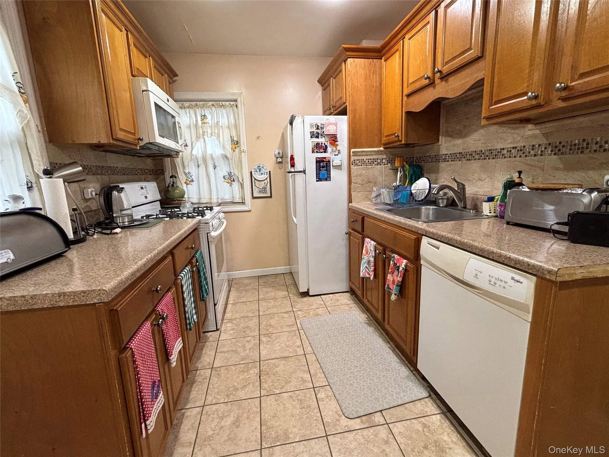 Kitchen with white appliances, backsplash, light tile patterned floors, and brown cabinetry Kitchen with white appliances, backsplash, light tile patterned floors, and brown cabinetry