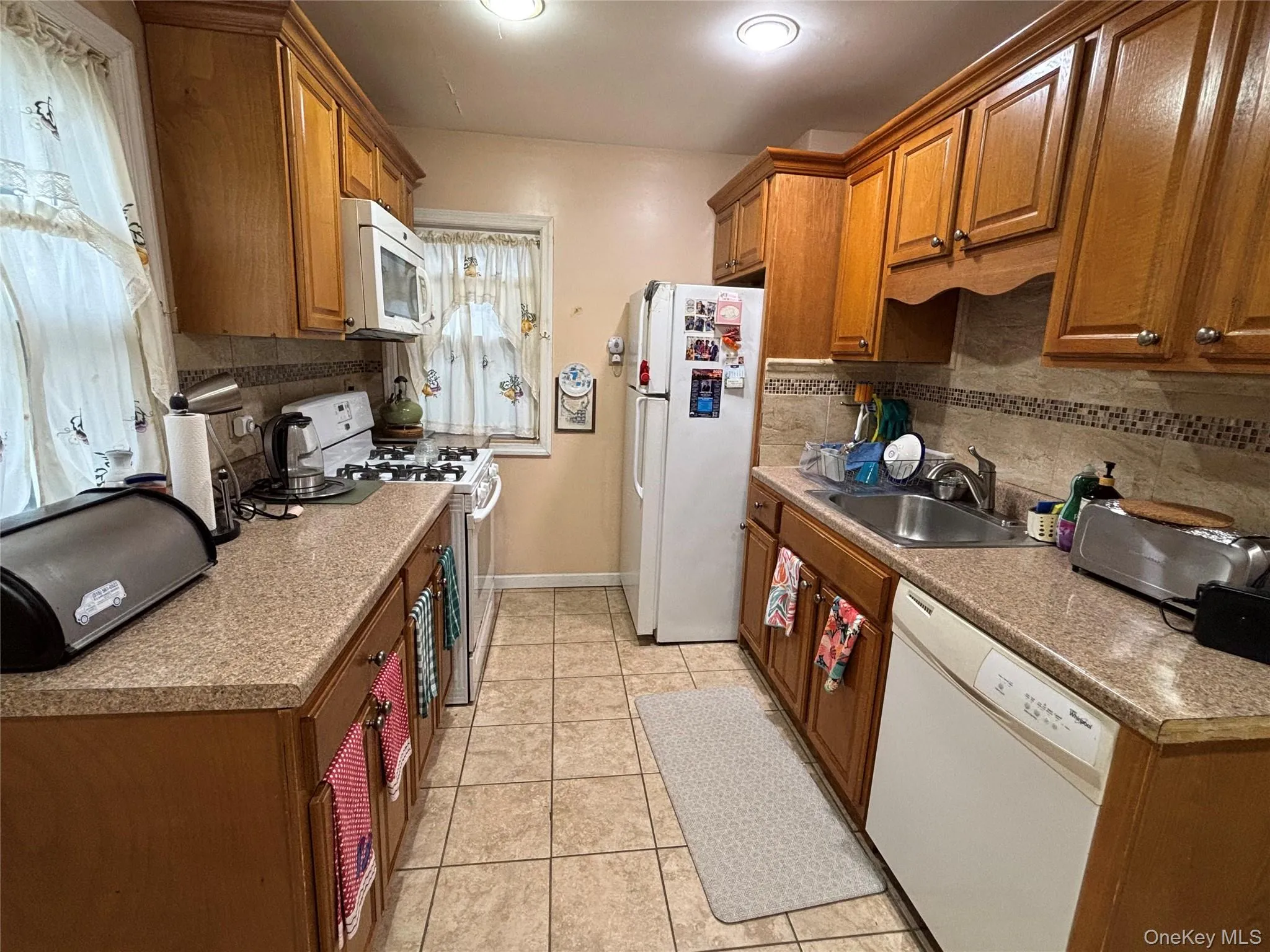 Kitchen featuring brown cabinets, white appliances, light tile patterned flooring, and backsplash Kitchen featuring brown cabinets, white appliances, light tile patterned flooring, and backsplash