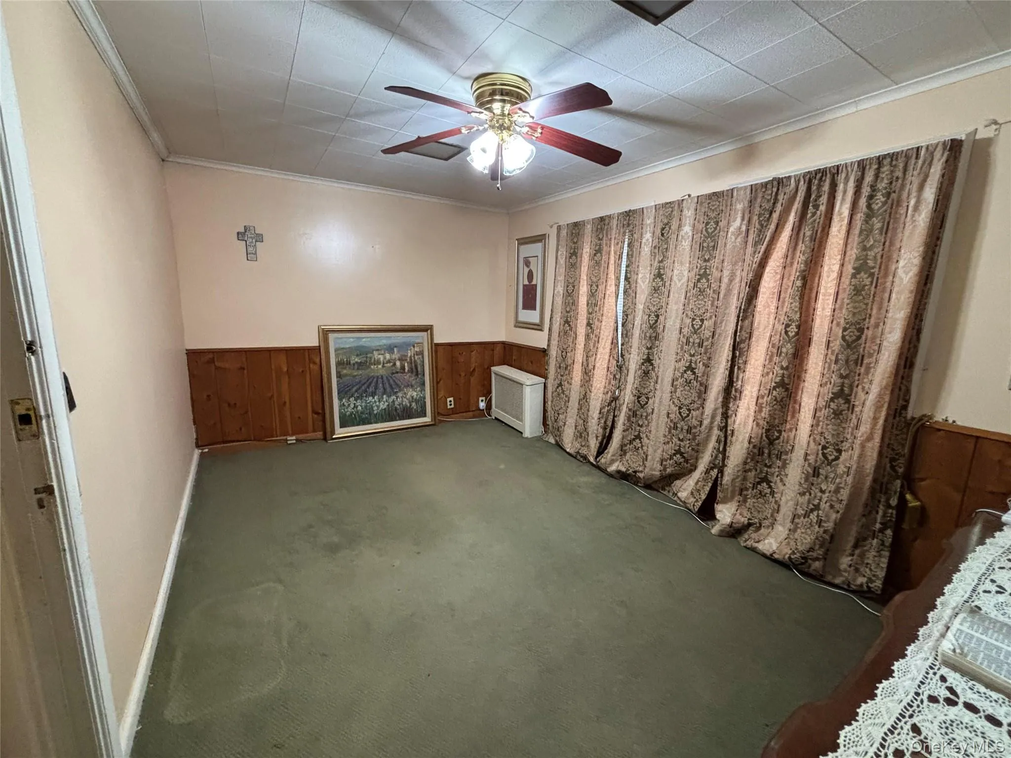 Carpeted empty room featuring wood walls, ornamental molding, wainscoting, ceiling fan, and radiator heating unit Carpeted empty room featuring wood walls, ornamental molding, wainscoting, ceiling fan, and radiator heating unit