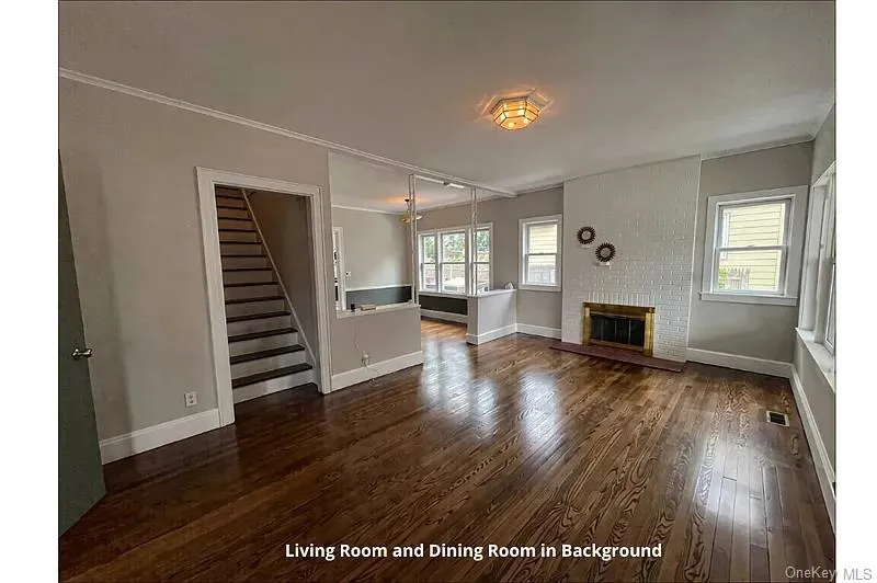 Unfurnished living room with dark wood-style flooring, a brick fireplace, stairway, healthy amount of natural light, and ornamental molding Unfurnished living room with dark wood-style flooring, a brick fireplace, stairway, healthy amount of natural light, and ornamental molding