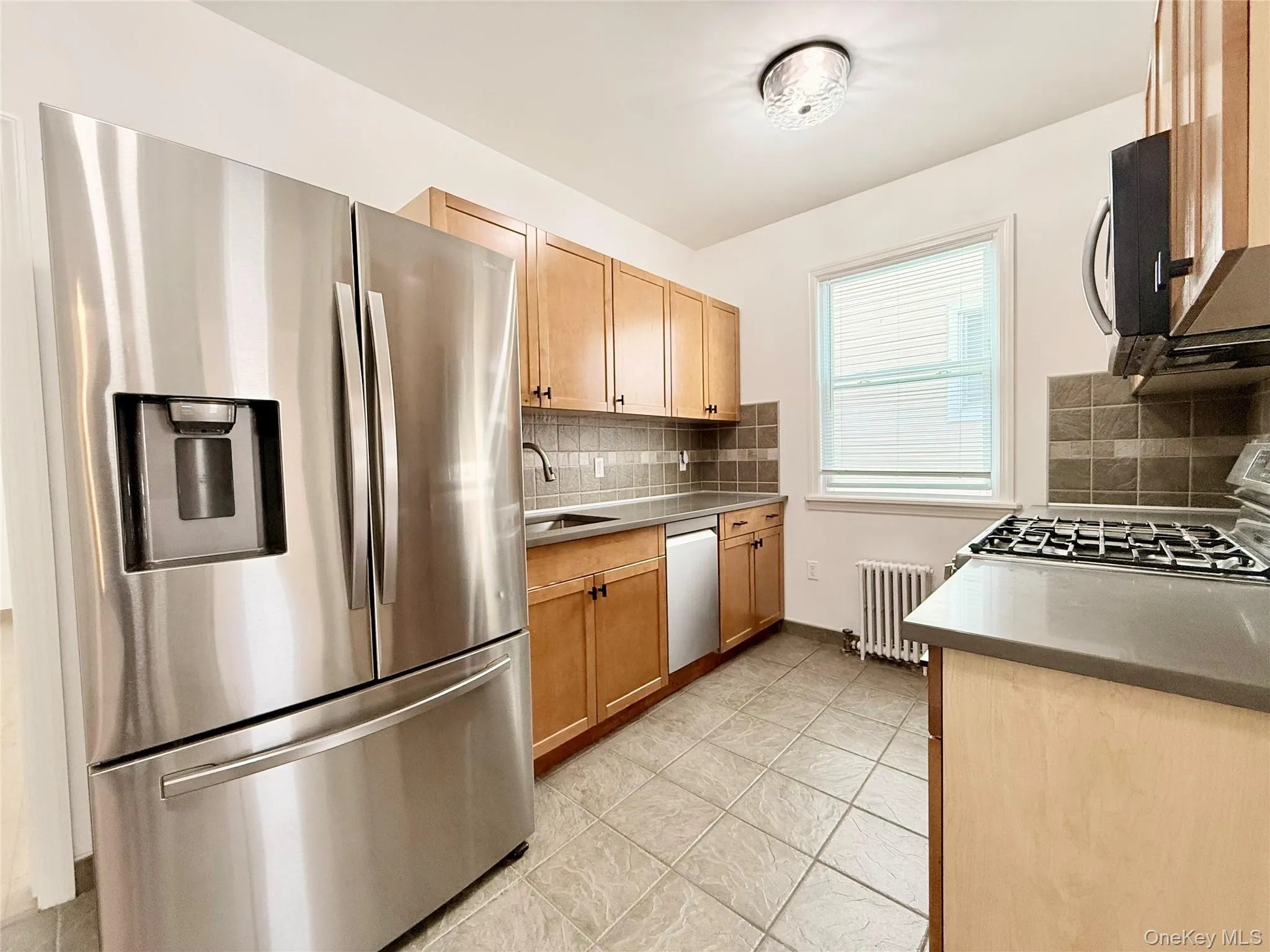 Kitchen featuring appliances with stainless steel finishes, tasteful backsplash, radiator heating unit, light tile patterned flooring, and light brown cabinets Kitchen featuring appliances with stainless steel finishes, tasteful backsplash, radiator heating unit, light tile patterned flooring, and light brown cabinets