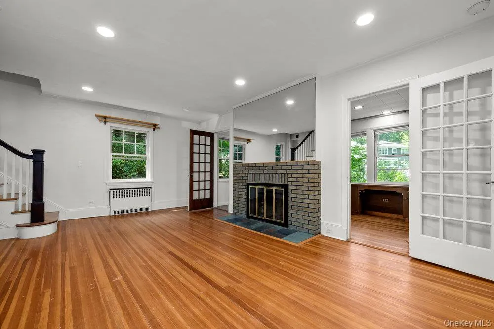 Unfurnished living room featuring stairs, recessed lighting, wood finished floors, radiator, and a brick fireplace Unfurnished living room featuring stairs, recessed lighting, wood finished floors, radiator, and a brick fireplace