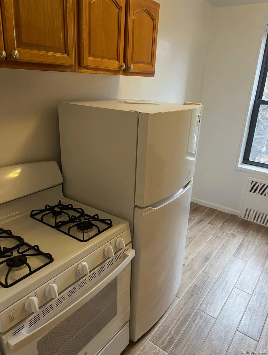 Kitchen featuring white range with gas cooktop, wood finish floors, brown cabinetry, radiator, and freestanding refrigerator Kitchen featuring white range with gas cooktop, wood finish floors, brown cabinetry, radiator, and freestanding refrigerator