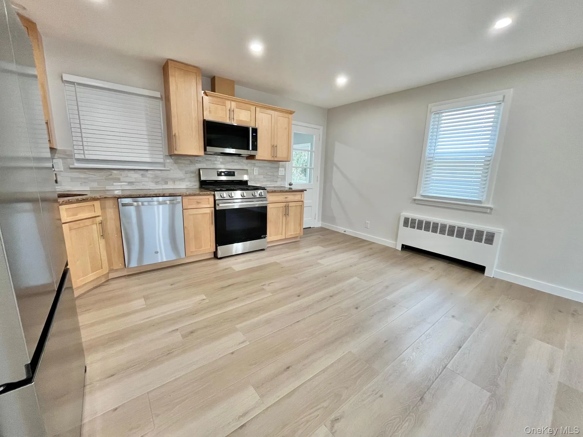 Kitchen with light brown cabinetry, stainless steel appliances, decorative backsplash, radiator, and light wood finished floors Kitchen with light brown cabinetry, stainless steel appliances, decorative backsplash, radiator, and light wood finished floors