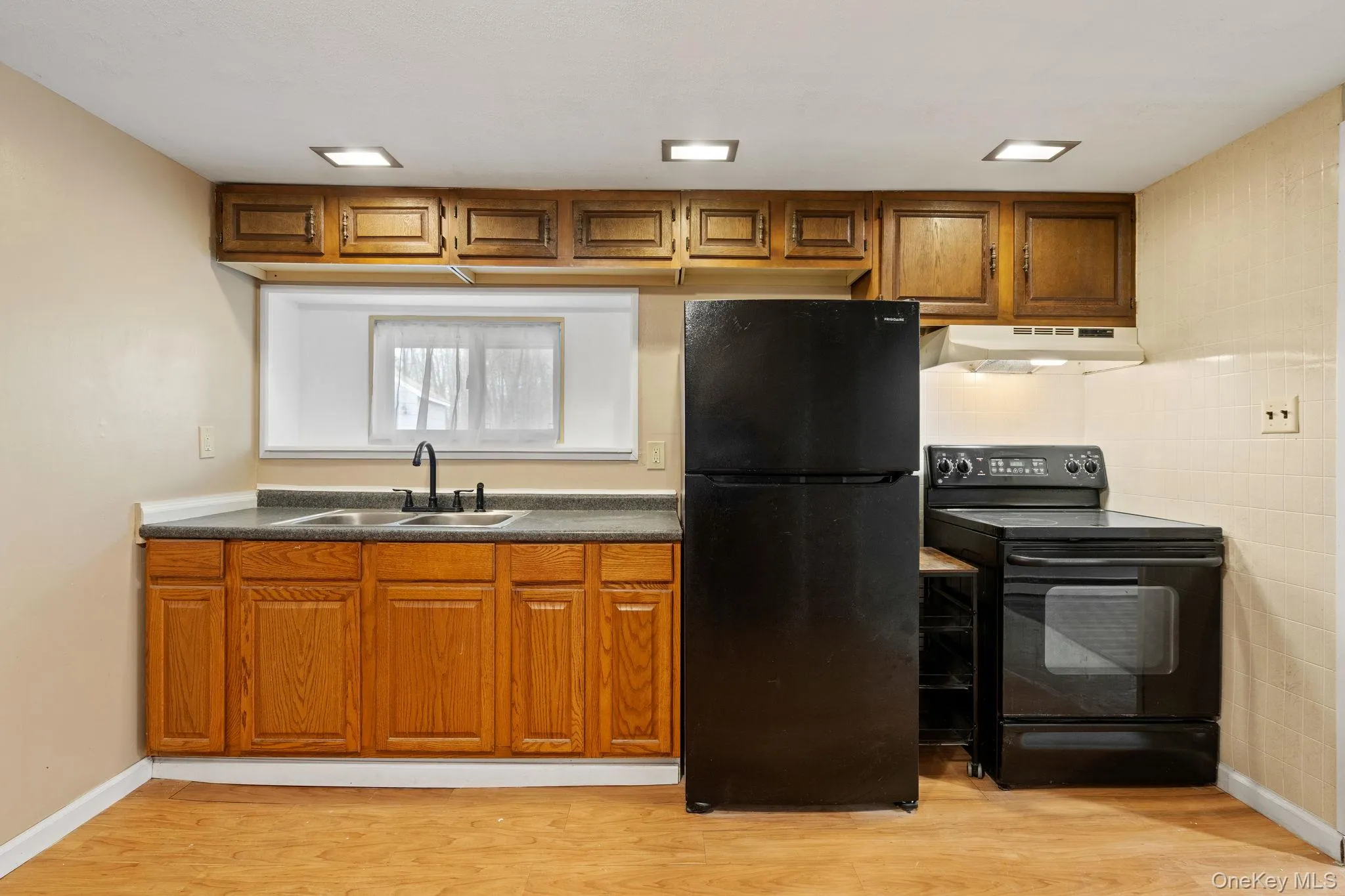 Kitchen with black appliances, brown cabinetry, dark countertops, and light wood-type flooring Kitchen with black appliances, brown cabinetry, dark countertops, and light wood-type flooring