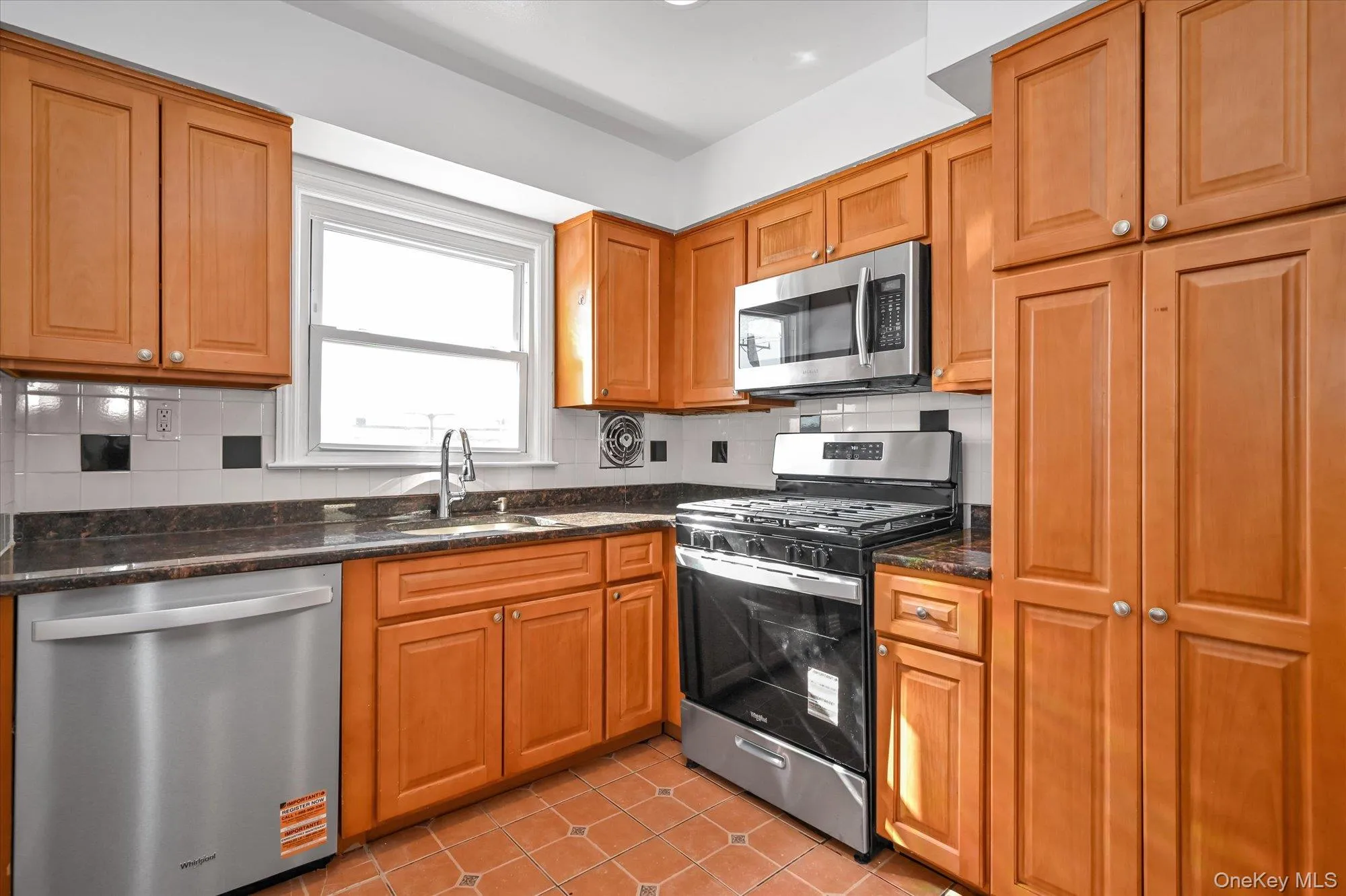 Kitchen featuring stainless steel appliances, backsplash, light tile patterned floors, dark stone counters, and brown cabinets Kitchen featuring stainless steel appliances, backsplash, light tile patterned floors, dark stone counters, and brown cabinets