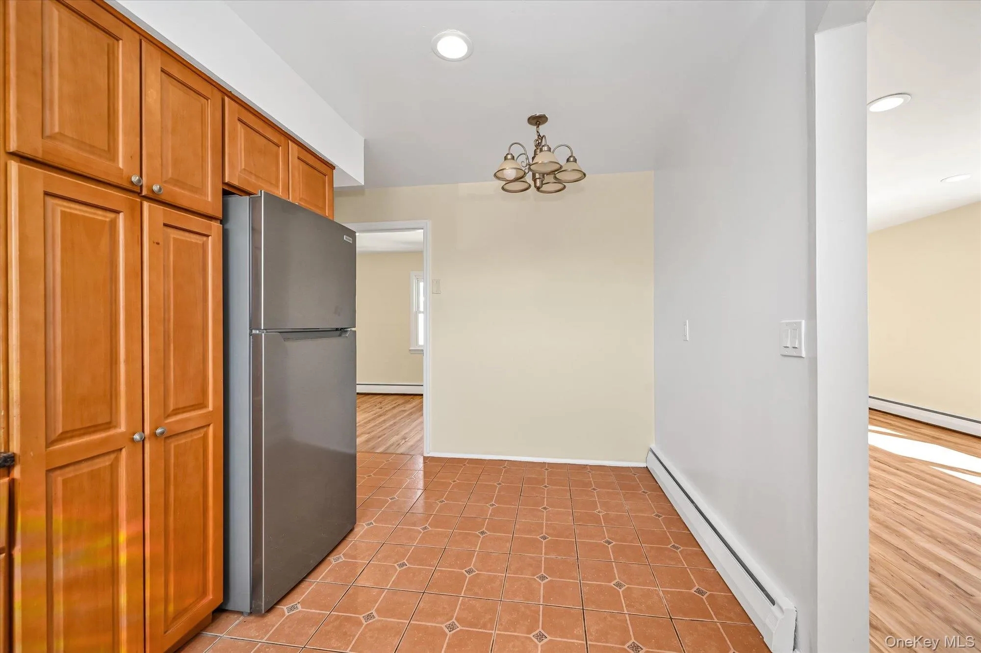 Kitchen featuring a baseboard radiator, freestanding refrigerator, brown cabinets, light tile patterned floors, and a chandelier Kitchen featuring a baseboard radiator, freestanding refrigerator, brown cabinets, light tile patterned floors, and a chandelier