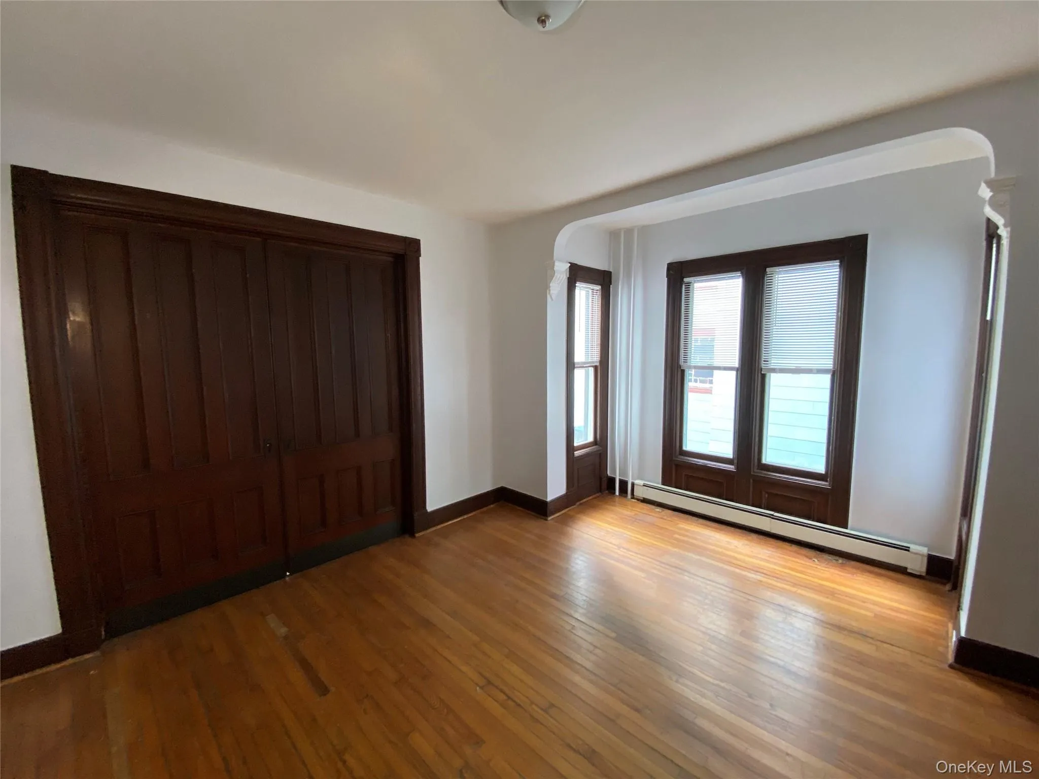 Unfurnished bedroom featuring baseboard heating, light wood-style flooring, arched walkways, and a closet Unfurnished bedroom featuring baseboard heating, light wood-style flooring, arched walkways, and a closet